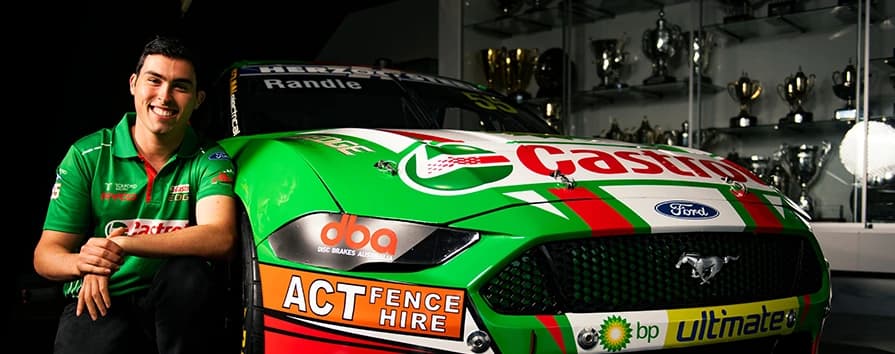 Pilote de course souriant en chemise verte d'équipe à côté d'une Ford Mustang de course verte sponsorisée par DBA avec logos de sponsors dans une salle de trophées